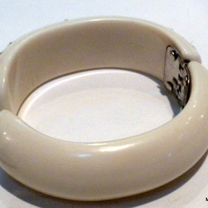 May include: Ivory-colored hinged bangle bracelet. The bracelet has a rounded shape with a smooth, glossy finish. Small, sparkling embellishments are set along the top edge of the bracelet. The clasp is silver-toned.