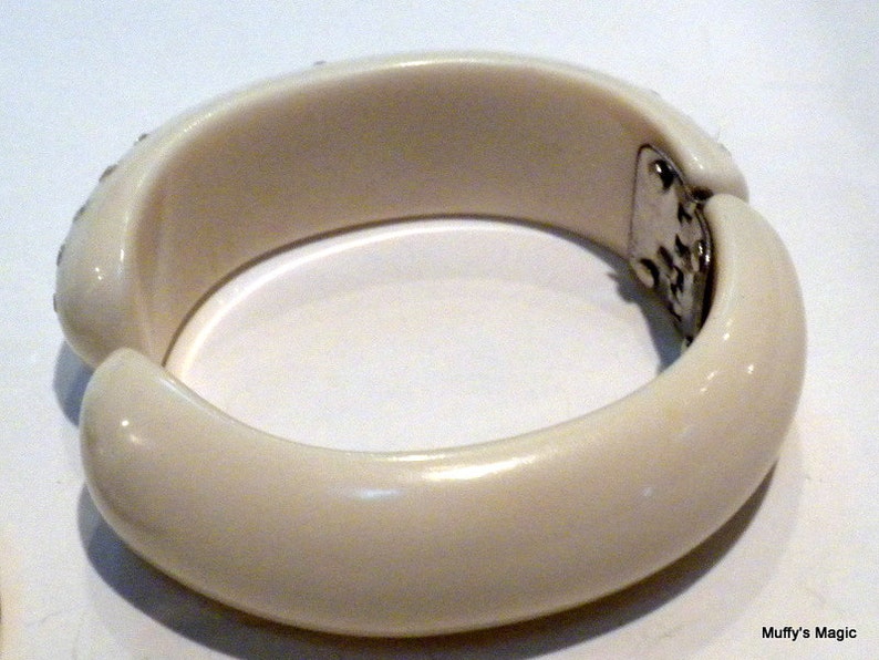 May include: Ivory-colored hinged bangle bracelet. The bracelet has a rounded shape with a smooth, glossy finish. Small, sparkling embellishments are set along the top edge of the bracelet. The clasp is silver-toned.