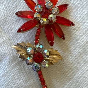 Könnte beinhalten: Vintage Blumenbrosche mit einer roten Edelsteinblume und Stiel, akzentuiert mit schillernden Strasssteinen und goldfarbenen Blättern. Die Brosche hat ein detailliertes Design mit einem klassischen, eleganten Stil.