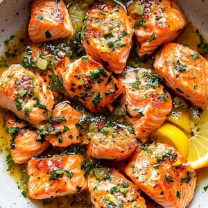 May include: A white bowl filled with pan-seared salmon pieces in a lemon butter sauce with fresh parsley and lemon slices.