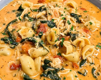 May include: A bowl of creamy tomato soup with orecchiette pasta, shredded chicken, spinach, and diced tomatoes. The soup is garnished with fresh parsley and served in a light blue bowl.