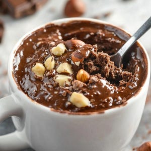 May include: A white mug filled with chocolate cake, topped with chopped hazelnuts and white chocolate pieces. A spoon is inserted into the cake, and chocolate pieces and whole hazelnuts are scattered around the mug.