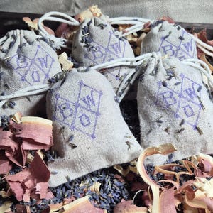 May include: Five small, beige drawstring bags with a purple geometric design and the letters "B W O". The bags are filled with dried lavender and sit among wood shavings and dried flowers. The bags are tied with white string.