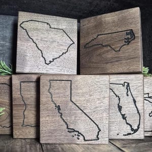 Puede incluir: Un conjunto de posavasos de madera con contornos grabados de estados de EE. UU. Los posavasos cuadrados tienen un acabado de veta de madera oscura. Se ven los estados de Carolina del Sur, Carolina del Norte, California, Florida y Michigan.