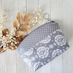 May include: A gray and white fabric zippered pouch with a white polka dot pattern on the top half and a white damask pattern on the bottom half. The pouch has a silver zipper pull with a ring.