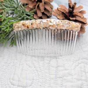 Op de afbeelding: Een doorzichtige plastic haar kam met een decoratieve bovenkant gemaakt van kleine, iriserende schelpstukken. De kam staat tegen een witte, getextureerde achtergrond met dennenappels en groen.