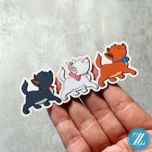 May include: Three cartoon cat stickers. One is black with a red bow, the second is white with a pink bow, and the third is orange with a blue bow. All three cats are looking upwards with happy expressions.