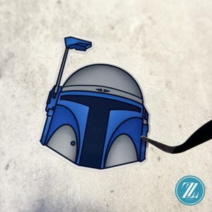 May include: A die-cut sticker featuring a stylized helmet in shades of blue, dark blue, and light gray. The helmet design includes a T-shaped visor, a prominent antenna extending from the left side, and a small circular detail on the lower right side.