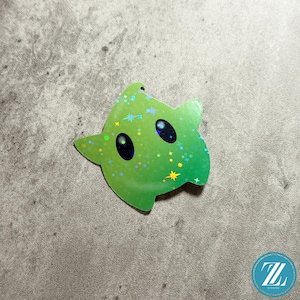 May include: A green star-shaped sticker with a holographic finish. The sticker features two large black eyes and small star accents. The sticker has a hole at the top for hanging. The sticker is on a gray surface.