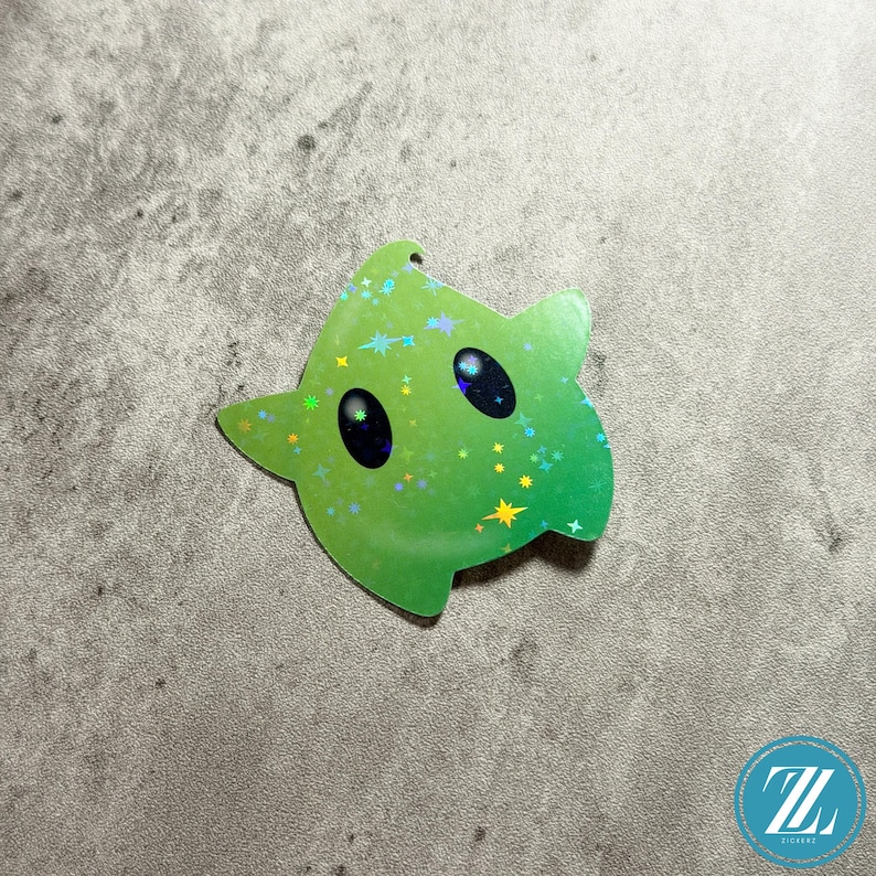 May include: A green star-shaped sticker with a holographic finish. The sticker features two large black eyes and small star accents. The sticker has a hole at the top for hanging. The sticker is on a gray surface.