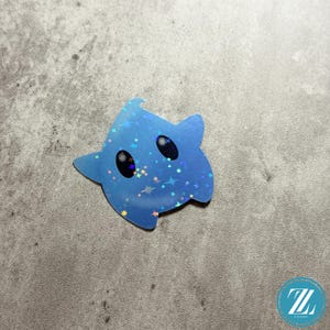 May include: A blue star-shaped sticker with a holographic finish. The sticker features two black oval eyes and a scattering of small stars. The sticker is on a gray surface.