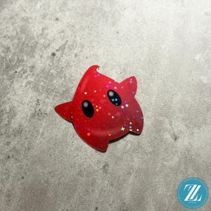 May include: A red star-shaped sticker with black oval eyes and a glittery, holographic finish. The sticker has a cartoonish design with a gradient effect, and is likely a decorative item or a collectible.