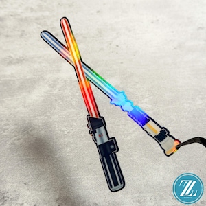 May include: A sticker featuring two crossed lightsabers. One lightsaber has a red blade and a black hilt, while the other has a blue blade and a silver and blue hilt. Both blades exhibit a holographic, iridescent sheen that reflects light in a spectrum of colors.