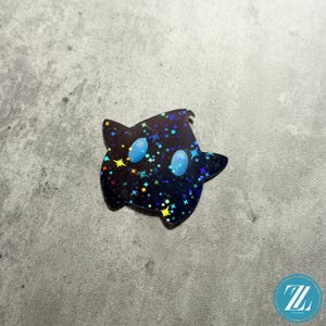 May include: A black sticker in the shape of a star with a cartoon ghost-like face. The sticker has a holographic finish with colorful stars and two light blue oval eyes. The sticker is on a gray surface.