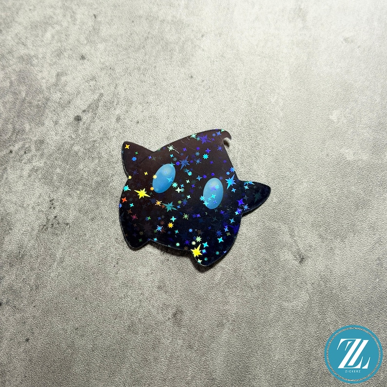 May include: A black sticker in the shape of a star with a cartoon ghost-like face. The sticker has a holographic finish with colorful stars and two light blue oval eyes. The sticker is on a gray surface.