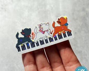 Puede incluir: Una pegatina con tres gatitos de dibujos animados tocando el piano. Los gatitos son negros, blancos y naranjas, cada uno con un lazo. Las teclas del piano son blancas y negras, con detalles brillantes. La pegatina se sostiene en una mano.