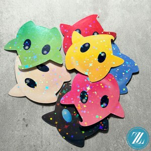 May include: A collection of colorful, star-shaped stickers with a cartoonish design. The stickers are in various colors, including green, red, yellow, blue, pink, and black, and feature a holographic, glittery finish. Each sticker has two black oval eyes.