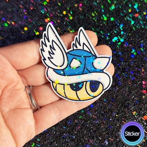 May include: A blue and white sticker featuring a cartoon turtle with wings. The sticker has a shiny, holographic finish with star-shaped accents. The sticker has a white border and a yellow underside. The word "Sticker" is in a circle.