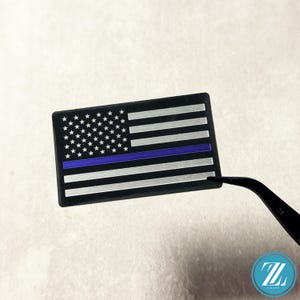 May include: A rectangular patch featuring a stylized American flag design in black and silver with a single horizontal blue stripe running through the center. The flag includes a field of stars and alternating horizontal stripes, held by black tweezers.