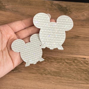 May include: Two white holographic stickers shaped like mouse heads with a geometric pyramid texture. Each sticker features a reflective surface that catches light to create small rainbow sparkles across the repeating triangular pattern.