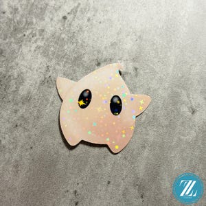 May include: A light pink sticker in the shape of a star-like character with large black eyes and a yellow star in one eye. The sticker is covered in iridescent stars.
