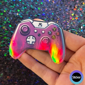 May include: A holographic sticker of an Xbox controller. The controller is primarily purple with rainbow accents and features the standard button layout. The sticker has a glossy finish and a white outline.