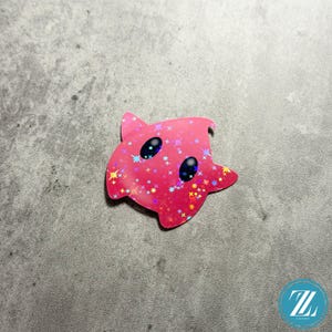 May include: A pink, star-shaped sticker with a holographic finish. The sticker features two black oval eyes and is covered in colorful star-shaped sparkles. The sticker is on a gray surface.