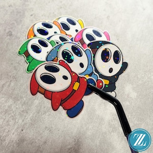 May include: A collection of colorful, holographic stickers featuring a cartoon character with a white mask and various colored outfits. The stickers are in shades of red, blue, green, orange, pink, and black. A pair of black tweezers holds one sticker.