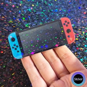 May include: A holographic sticker of a Nintendo Switch console with a black background and colorful stars. The sticker is designed to look like a real Nintendo Switch console.