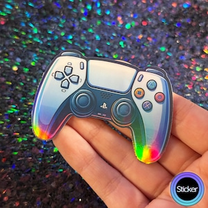May include: A holographic sticker of a white PlayStation 5 controller with black buttons and a rainbow iridescent finish. The sticker is on a hand.