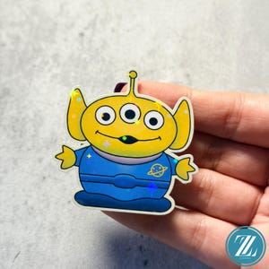 May include: A holographic sticker featuring a cartoon alien character with three eyes, yellow head and ears, and a blue uniform. The sticker has a shiny, iridescent finish and is held in a hand.