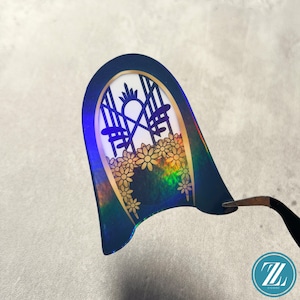 May include: A navy blue and gold sticker with a floral and geometric design. The sticker has a holographic effect, with a window-like design featuring flowers and a sun. The sticker is being held by tweezers.