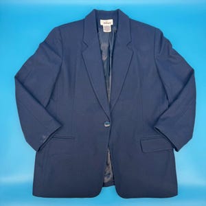 May include: A solid navy blue blazer featuring a single-button closure, notched lapels, and two front flap pockets. The interior is lined with a matching dark fabric, and a white brand label is visible at the center of the inner neckline.