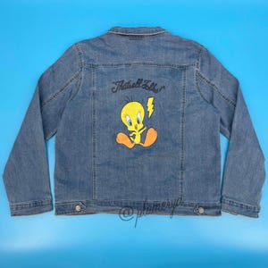 May include: A blue denim jacket featuring a graphic on the back of the yellow cartoon character Tweety Bird with orange feet and a yellow lightning bolt. Above the character, the text reads That's all folks! in a black cursive script font.