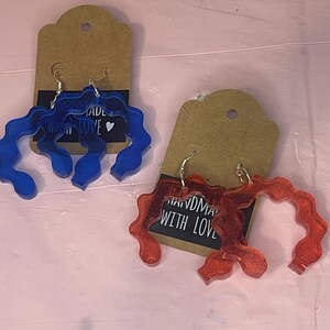 May include: Two pairs of handmade earrings. One pair is blue and the other pair is red with glitter. Both pairs are made in the shape of a wavy line.