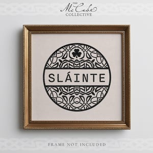 May include: A square framed print featuring a black circular design with intricate Celtic knotwork and a shamrock at the top. The word SL&Aacute;INTE is printed in black capital letters across a horizontal bar in the center of the circle. The frame is gold.