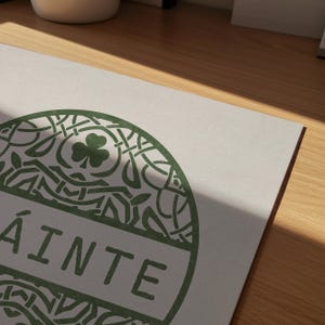 May include: A rectangular white print featuring a circular green graphic with intricate Celtic knotwork patterns, a central shamrock icon, and the word SL&Aacute;INTE printed in bold green capital letters across the middle of the circular design.