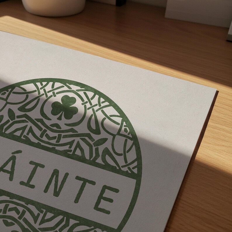May include: A rectangular white print featuring a circular green graphic with intricate Celtic knotwork patterns, a central shamrock icon, and the word SL&Aacute;INTE printed in bold green capital letters across the middle of the circular design.