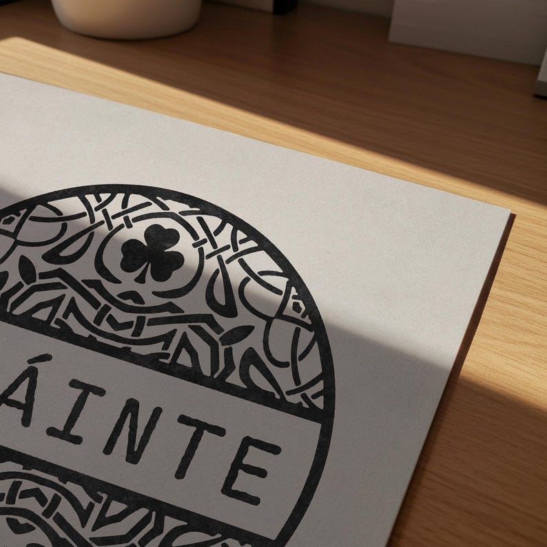 May include: A rectangular white card featuring a circular black graphic with intricate Celtic knotwork patterns, a central shamrock icon, and the word "SL&Aacute;INTE" printed in bold black capital letters across the center of the circular design.