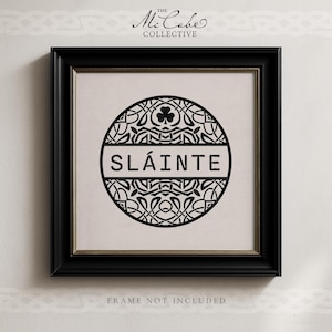 May include: A square black frame containing a white print with a circular black design featuring intricate knotwork, a shamrock icon, and the text SL&Aacute;INTE in the center. The text THE MCCABE COLLECTIVE appears above the frame and FRAME NOT INCLUDED below.