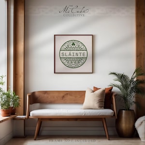 May include: A square wall art print featuring a circular green Celtic knot design with a shamrock at the top and the text SL&Aacute;INTE in the center. The print is displayed above a wooden bench with a white cushion and two throw pillows.