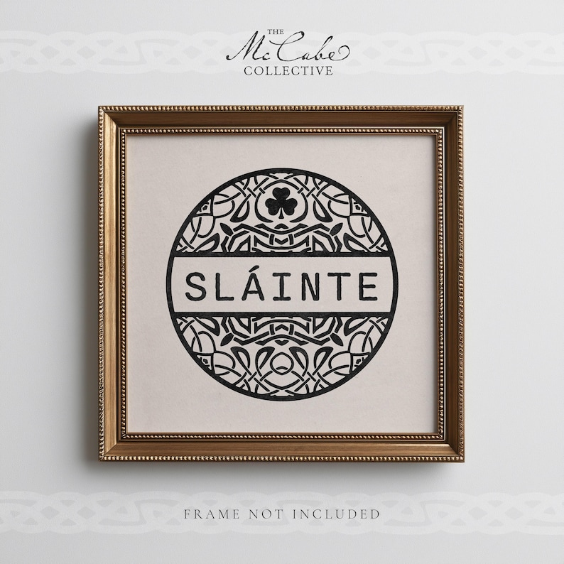 May include: A square framed print featuring a black circular design with intricate Celtic knotwork and a shamrock at the top. The word SL&Aacute;INTE is printed in black capital letters across a horizontal bar in the center of the circle. The frame is gold.