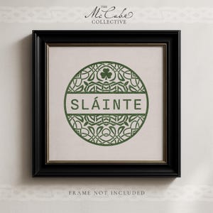 May include: A square black frame containing a print with a circular green Celtic knot design. The center features the word SL&Aacute;INTE in green capital letters. Above the text is a small shamrock. The text THE MCCABE COLLECTIVE is at the top.