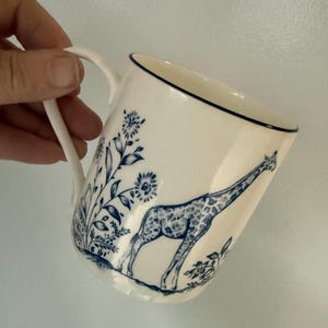 May include: White ceramic mug with a blue giraffe illustration and floral accents. The mug has a curved handle and a dark blue trim along the rim. The giraffe is depicted with a spotted pattern.