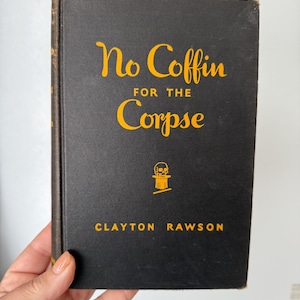 Może przedstawiać: Vintage książka w twardej oprawie zatytułowana "No Coffin for the Corpse" autorstwa Claytona Rawsona. Książka ma czarną okładkę ze złotymi literami i małą ilustracją czaszki. Książka jest trzymana przez osobę.