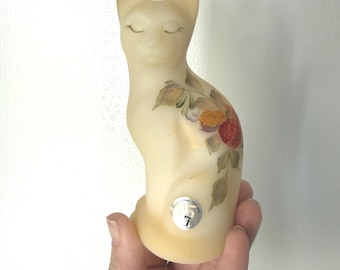Peut inclure: Figurine de chat crème avec les yeux fermés et des détails floraux peints. Le chat a un autocollant sur sa base. Les fleurs sont rouges, jaunes et blanches avec des feuilles vertes. La figurine est tenue dans une main.