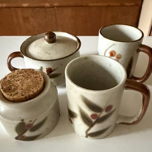 May include: A ceramic tea set featuring a sugar bowl with a cork lid, a lidded creamer, and two mugs. The set has a speckled, off-white glaze with brown handles and decorative leaf and berry motifs. The mugs are approximately 4 inches tall.