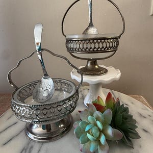 May include: Two antique silver-plated serving dishes with spoons. One dish has a handle, and both have intricate openwork designs. A small white cake stand and a cluster of faux succulents in shades of green, blue, and red are also present.