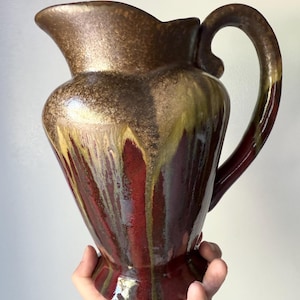 May include: A ceramic pitcher featuring a glossy glaze with a deep burgundy base color and a metallic bronze or gold drip pattern cascading from the rim. The vessel has a wide spout, a curved handle, and a flared pedestal base with a smooth finish.