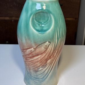 May include: A ceramic vase with a teal and peach color scheme. The vase features a circular indentation and a relief design of a fish. The vase has a smooth, glossy finish and a unique, artistic shape.